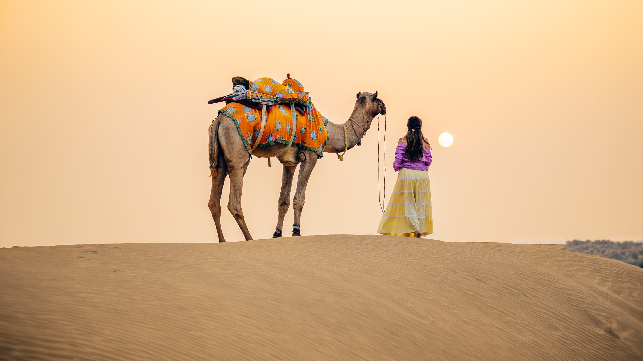 Women wearing dress in the desert with a camel at sunset